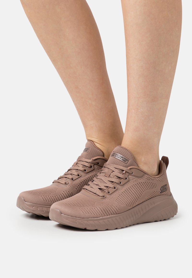 Pieds portant des chaussures de sport tricotées marron clair avec fermeture à lacets et semelles texturées sur fond blanc.
