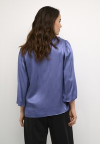 Blusa in satin di colore lilla con maniche a tre quarti e vestibilità morbida. Texture liscia con una leggera lucentezza. Abbinata a pantaloni neri.
