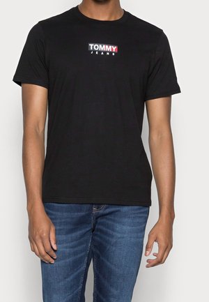 Mężczyzna w czarnej koszulce z krótkim rękawem z napisem "TOMMY JEANS" i niebieskich dżinsach, stojący na jednolitym tle.