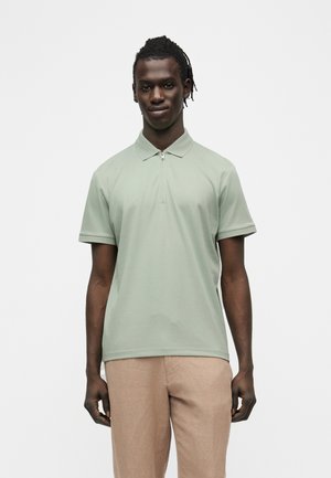 Dorosły mężczyzna stojący na białym tle, ubrany w jasnooliwkową koszulkę polo z krótkim rękawem i suwakiem oraz beżowe spodnie.