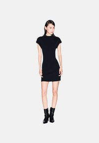 Abito aderente nero con maniche corte, scollatura rotonda e lunghezza fino al ginocchio, abbinato a stivali ankle con tacco neri. Tessuto liscio, design minimalista.