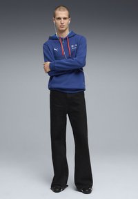 Blauer Kapuzenpullover mit rotem Kordelzug und Puma-Logo, kombiniert mit schwarzen Schlagjeans und schwarzen Schuhen. Weiße Ohrhörer sind um den Hals getragen.