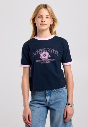 Tienermeisje met blond haar dat een marineblauw T-shirt met roze rand en de tekst "Bahamas Sports Department Surf Club" draagt, gecombineerd met lichtblauwe spijkerbroek.