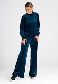 Zestaw dresowy z aksamitnego materiału w głębokim teal, składający się z luźnej bluzki z podwyższonym kołnierzem i rozszerzanych spodni, ozdobiony szwami bocznymi i miękką teksturą.