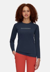 Γυναίκα με σγουρά μαλλιά φορώντας μπλε μακρυμάνικη μπλούζα με το κείμενο "MAMMUT" και φωτεινά πορτοκαλί παντελόνια, στέκεται με το ένα χέρι στην τσέπη.