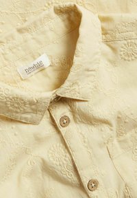 Gele katoenen shirt met complexe bloemenborduursels, knoopsluiting met houten knopen en een kraagontwerp. Beperkte oplage label zichtbaar.