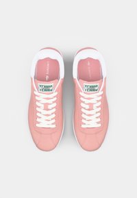 Ροζ υφασμάτινα αθλητικά παπούτσια με λευκές καουτσούκ σόλες, λευκές κορδόνες και πράσινες ετικέτες που γράφουν "LACOSTE" και "TENNIS". Στρογγυλή μύτη και επενδεδυμένος γιακάς.