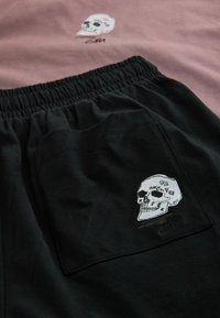 Un short noir avec une taille élastique comporte une poche latérale ornée d'une broderie blanche représentant un crâne. Au-dessus, une chemise rose affiche un motif de crâne similaire.