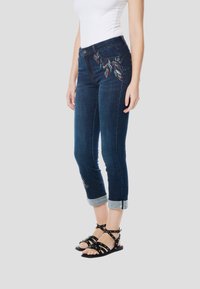 Sötétkék farmernadrág, hímzett levélmintával a csípőn, felhajtott szegélyekkel és közepes derékmagassággal. Fekete pántos szandállal párosítva.