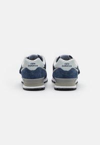 Baskets en daim bleu marine avec accents blancs, arborant une finition texturée, un col rembourré et un logo New Balance réfléchissant au talon.