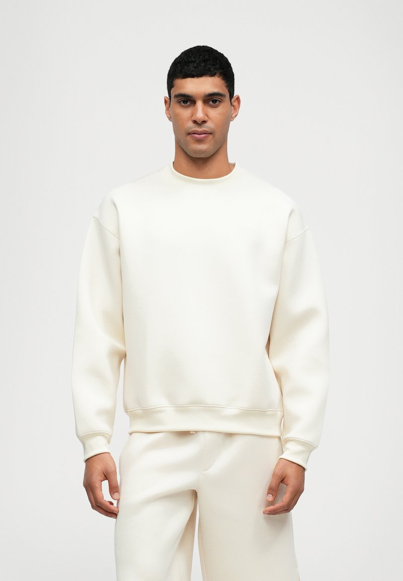 Sweatshirt crème avec une coupe décontractée, des poignets et un ourlet côtelés, fabriqué en tissu lisse. Design minimaliste sans logos visibles.