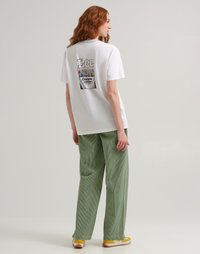 Donna con capelli rossi che indossa una t-shirt bianca Lee con un design di pastelli Crayola sulla schiena, pantaloni a righe verdi e sneakers gialle, in piedi di lato.