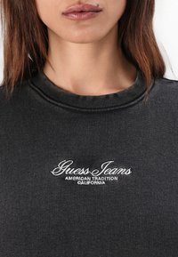 Melnā džemperis ar rievotu apkakli, priekšpusē ar baltu izšuvumu "Guess Jeans" un "AMERICAN TRADITION CALIFORNIA".