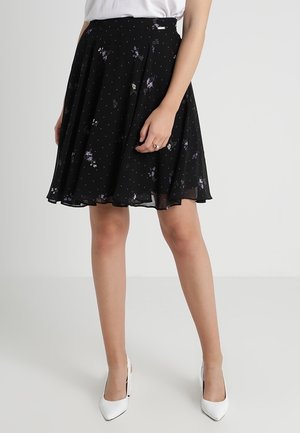 Frau trägt einen knielangen schwarzen Rock mit kleinen Blumenmustern, weiße spitze Absätze und ein weißes Oberteil, steht vor einem einfachen Hintergrund.