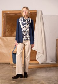 Mörkblå kofta över mönstrad blus, cremefärgade byxor och ankelboots med klack. Håller i en blå väska. Texturerad halsduk med mörkblått och cremefärgat mönster.