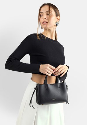 Donna che indossa un crop top nero a maniche lunghe e una gonna bianca, che tiene una piccola borsa in pelle nera con manici superiori contro uno sfondo semplice.