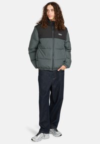 Persona con lunghi capelli ricci che indossa un giubbotto imbottito grigio e nero bicolore, jeans scuri e sneaker bianche, in piedi con le mani in tasca.