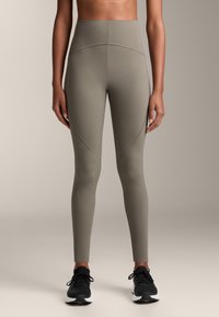 Magas derekú, olajzöld leggings rugalmas anyagból, oldalsó varrásokkal a kontúrozás érdekében, fekete sportcipővel párosítva.