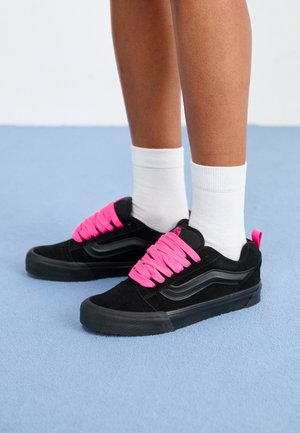 Persona con zapatillas negras con cordones rosa brillante y calcetines blancos hasta la pantorrilla, de pie sobre una alfombra azul claro.