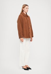 Giacca oversize in lana marrone con colletto, chiusura a bottoni e tessuto strutturato, abbinata a pantaloni a gamba larga color crema e tacchi neri.