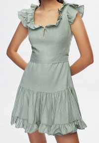 Helles grünes Leinenkleid mit Rüschensäumen, einem Schlüsselloch-Ausschnitt und einem gestuften, voluminösen Saum. Glatte Textur und taillierte Passform.