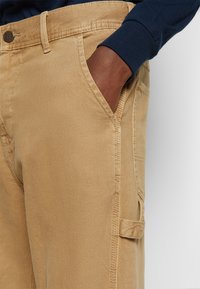 Pantalon cargo en coton kaki avec une poche latérale, des coutures renforcées, des passants pour ceinture et une fermeture à bouton. Texture lisse et coupe ajustée.