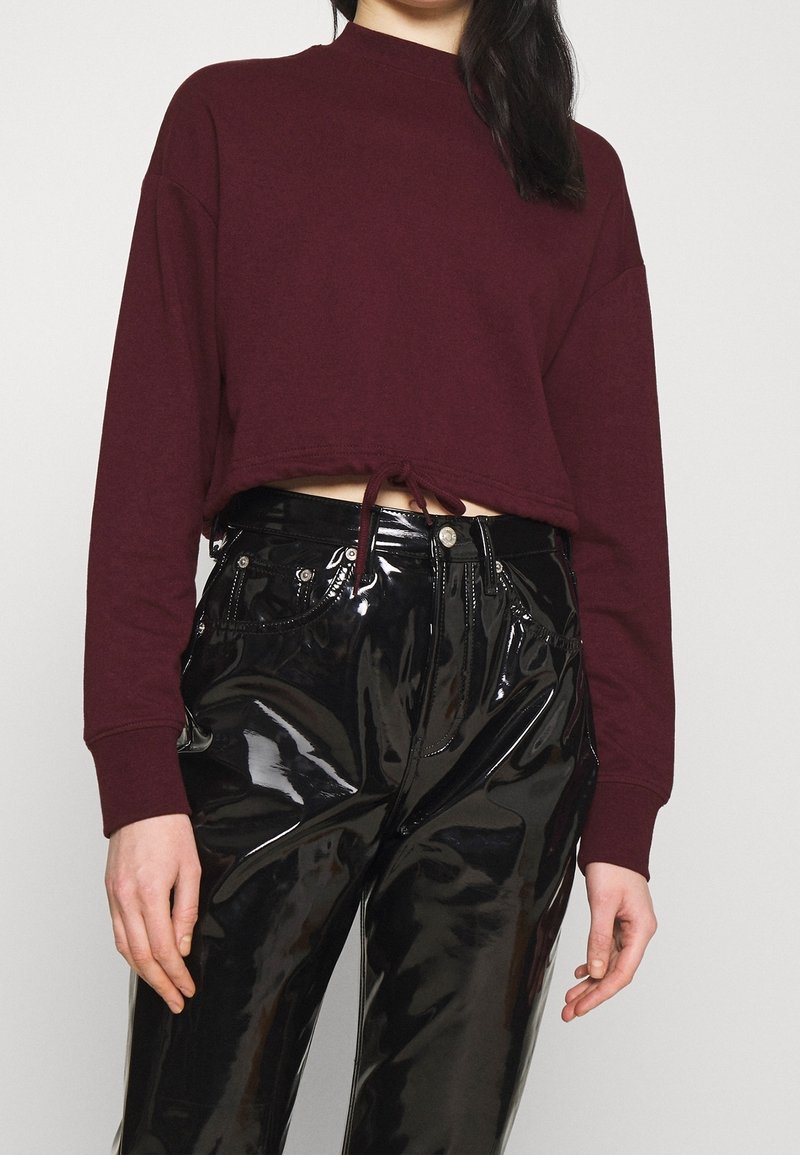 Personne portant un pull bordeaux court à manches longues et un pantalon en vinyle noir brillant taille haute sur un fond uni.