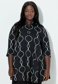 Donna sicura di sé che indossa una tunica nera con linee verticali bianche ondulate e maniche a tre quarti, in piedi contro uno sfondo bianco.