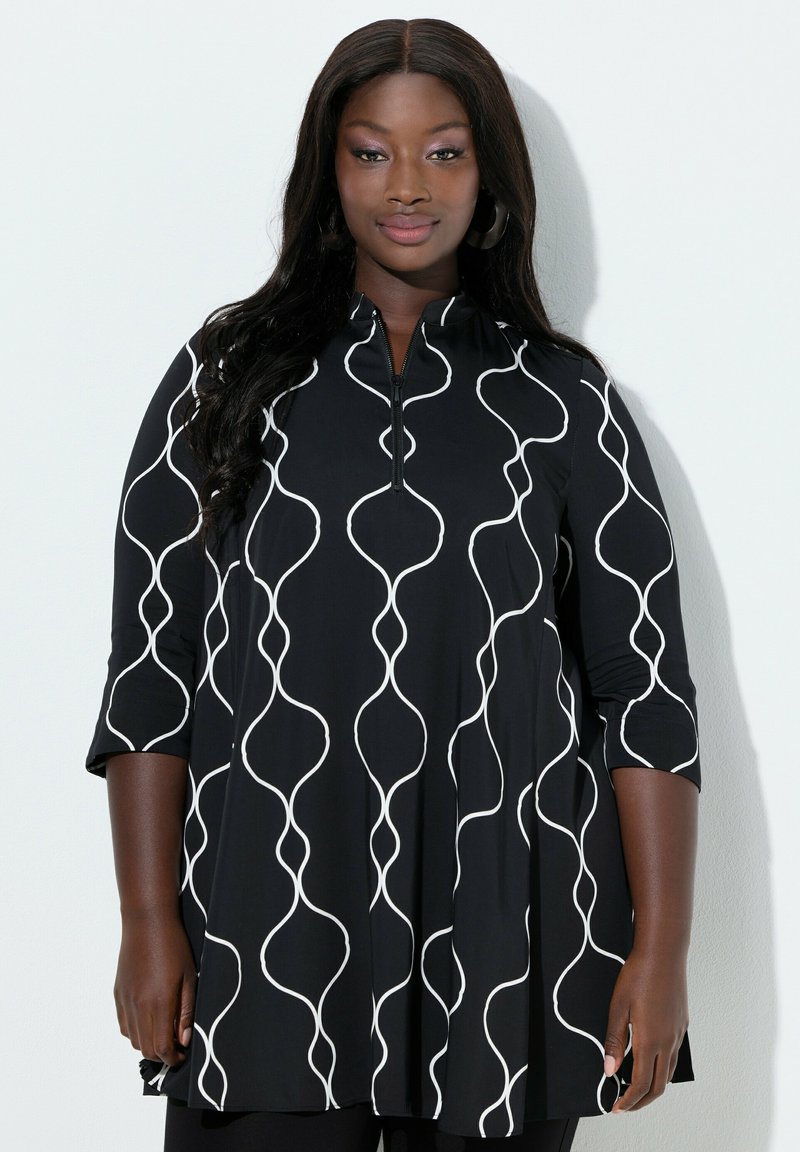 Donna sicura di sé che indossa una tunica nera con linee verticali bianche ondulate e maniche a tre quarti, in piedi contro uno sfondo bianco.