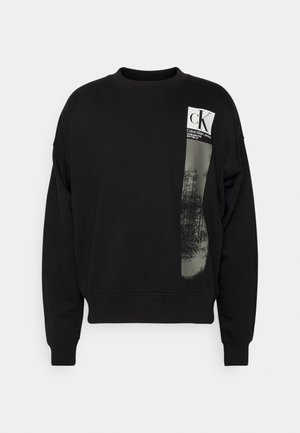 Czarna bluza z długim rękawem i okrągłym dekoltem, z naszywką z logo Calvin Klein Jeans i pionowym szarym teksturowanym paskiem na górnej lewej części przodu.