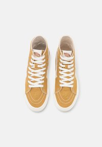 Höga sneakers i senapsgul canvas med vita snören, pryds av broderade sol- och blommönster nära tån och sidan, gummisulor.