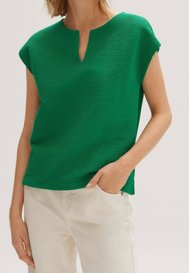 Zelený žebrovaný top s výstřihem do V, krátkými rukávy a texturovanou tkaninou. Kombinovaný s kalhotami světlé barvy, které ukazují neformální střih.