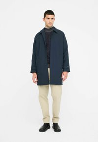 Cappotto impermeabile blu navy con chiusura a bottoni, tasche laterali e colletto. Indossato sopra un maglione scuro a trama e pantaloni beige. Stivali neri.