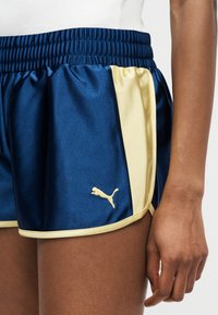 Κοντινό πλάνο ενός ατόμου που φοράει αθλητικές βερμούδες Puma σε ναυτικό μπλε και ανοιχτό κίτρινο με ελαστική μέση και λογότυπο στο αριστερό μπούτι.