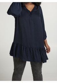 Mörkblå blus med trekvartslånga ärmar, V-ringning och knäppning; har en utsvängd, volangfåll och mjukt, texturerat tyg.