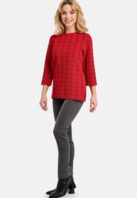 Rotes kariertes Oberteil mit dreiviertel Ärmeln, kombiniert mit grauen Slim-Fit-Hosen und schwarzen Stiefeletten, die strukturierte Muster betonen.