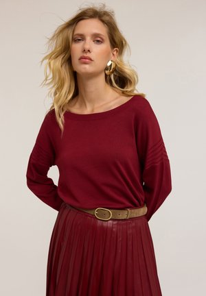 Bordeaux gebreide trui met lange mouwen, met geribbelde schouderdetails. Gecombineerd met een geplooide bordeaux rok en een bruine riem met gouden hardware.