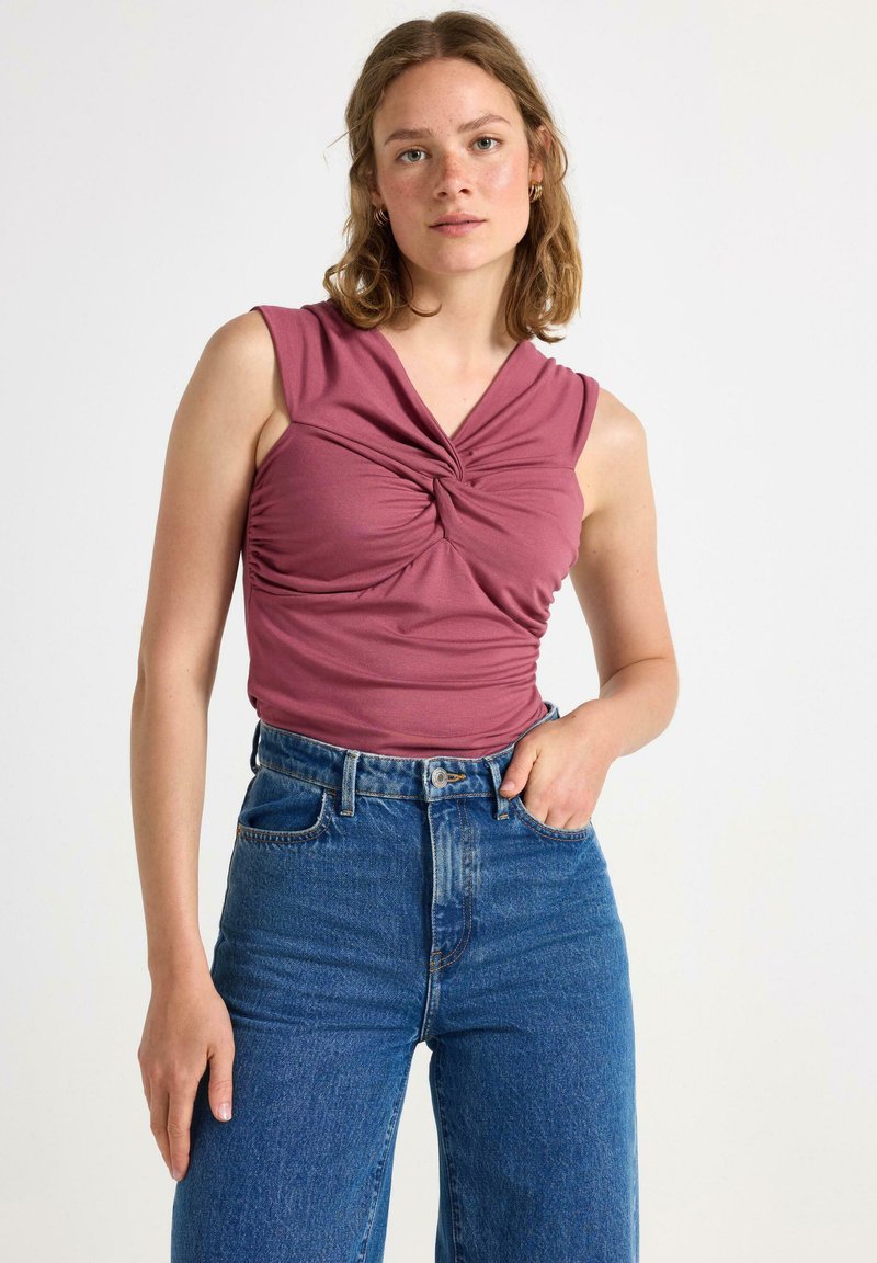 Lyhythihainen, kierretty etutoppi pehmeässä vaaleanpunaisessa kangassa; yhdistetty korkeavyötäröisiin, suoriin sinisiin farkkuihin, joissa on klassinen denimviimeistely.