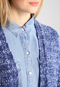 Blå stickad kardigan med vita prickar, med öppen front. Bärs över en ljusblå skjorta med knappar, krage och bröstficka.