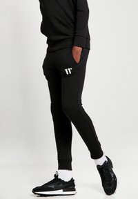 Pantalones de chándal deportivos negros, fabricados en tejido suave, con un diseño ajustado, que presentan un logo blanco '11°' en la parte frontal y bolsillos con puños acanalados.