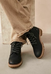 Botas de cuero negro con suelas robustas, con cordones y costuras en gris contrastante. Combinadas con pantalones beige claro, ligeramente desgastados.