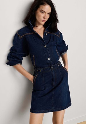 Donna che indossa un vestito di jeans scuro con maniche arrotolate, bottoni frontali e cuciture decorative, in piedi contro un muro bianco con le mani nelle tasche.