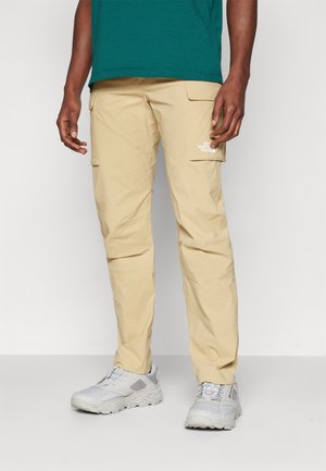 Jasnobeżowe spodnie cargo o zwężanym kroju, z bocznymi kieszeniami i detalem z logo. Noszone z szarymi sneakersami, prezentujące wielowarstwowy design.