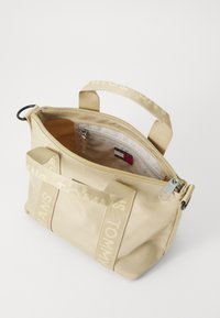 Beige stof håndtaske med dobbelte top-håndtag, lynlås lukning og en indvendig lomme. Har logo-stropper og en glat tekstur.