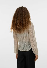 Genomskinlig, mönstrad blus med långa ärmar i beige spets. Har en rundad fåll och en strukturerad design med blommönster. Bärs tillsammans med svarta byxor.