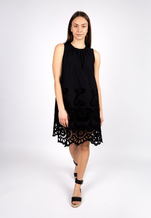 Schwarzes, ärmelloses Kleid mit aufwendigen Spitzenverzierungen am Saum und einem gerafften Ausschnitt. Sandalen mit schwarzen Riemen ergänzen das Outfit.