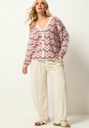 Donna con lunghi capelli biondi che indossa un cardigan bianco con motivi a zigzag rossi, pantaloni ampi color crema e sandali bianchi a fascia.