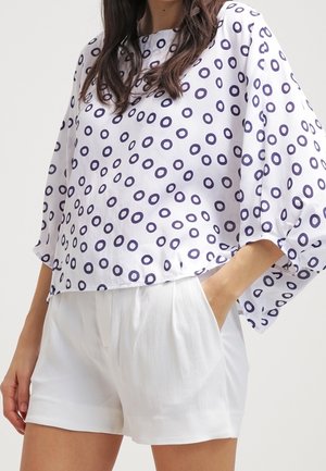 Kobieta nosząca luźną białą bluzkę z granatowymi okrągłymi wzorami oraz białe krótkie spodenki z wysokim stanem i kieszeniami, ręce w kieszeniach.