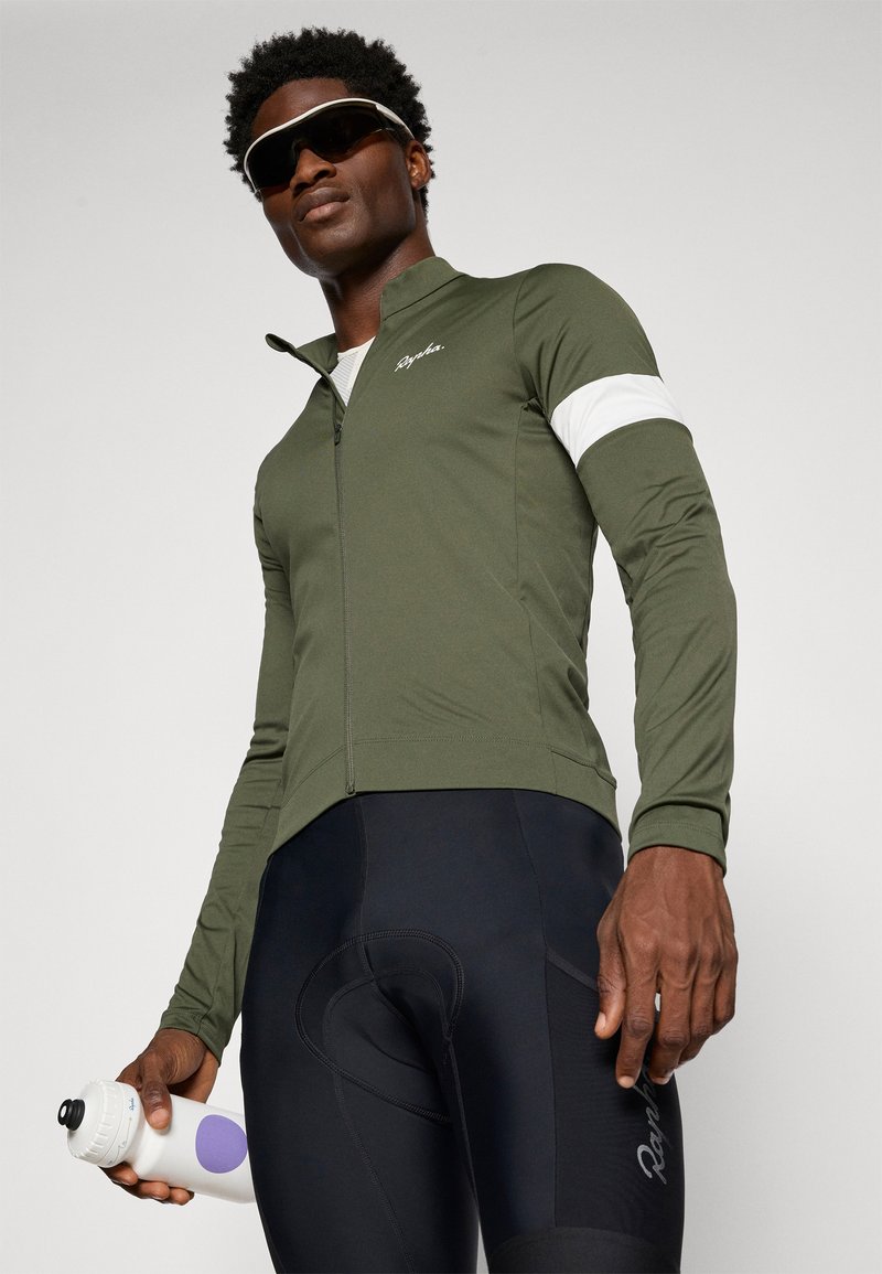 Mörkgrön zip-up cykeljacka med ett vitt rand på ärmen, tillsammans med svarta cykelshorts och håller en vit vattenflaska med lila detaljer.