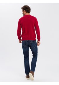 Czerwony sweter z dzianiny, z długimi rękawami, ściągaczami przy rękawach i u dołu, w zestawie z ciemnoniebieskimi dżinsami i brązowymi butami. Widoczny detal w postaci szwów na swetrze.